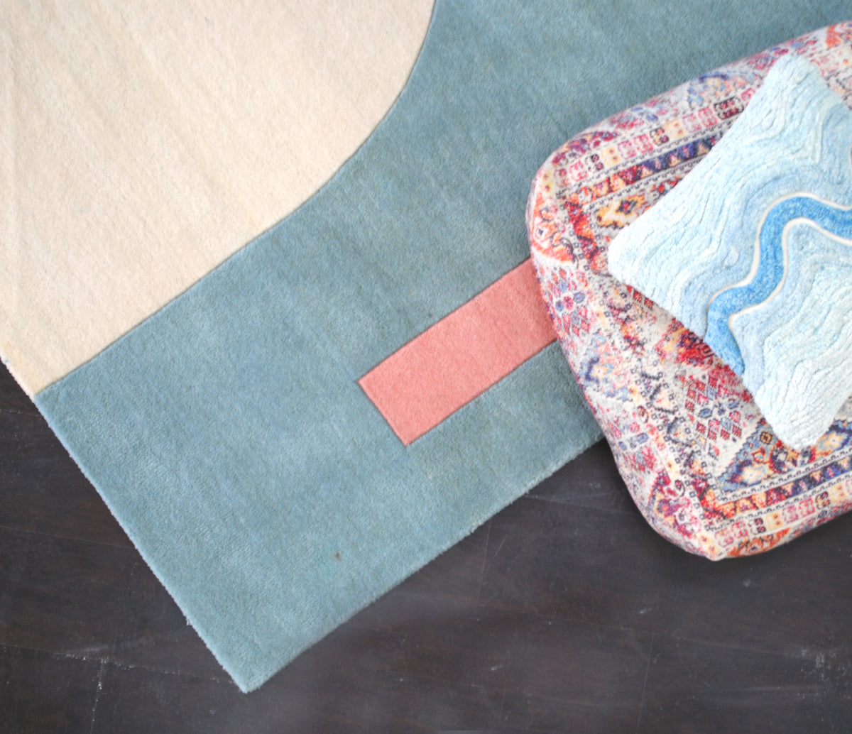 Corner of the Pondicherry hand-tufted wool rug from above, the coral bar running across the sea-glass blue field below the ivory arch, styled with a floor cushion, Art Avenue