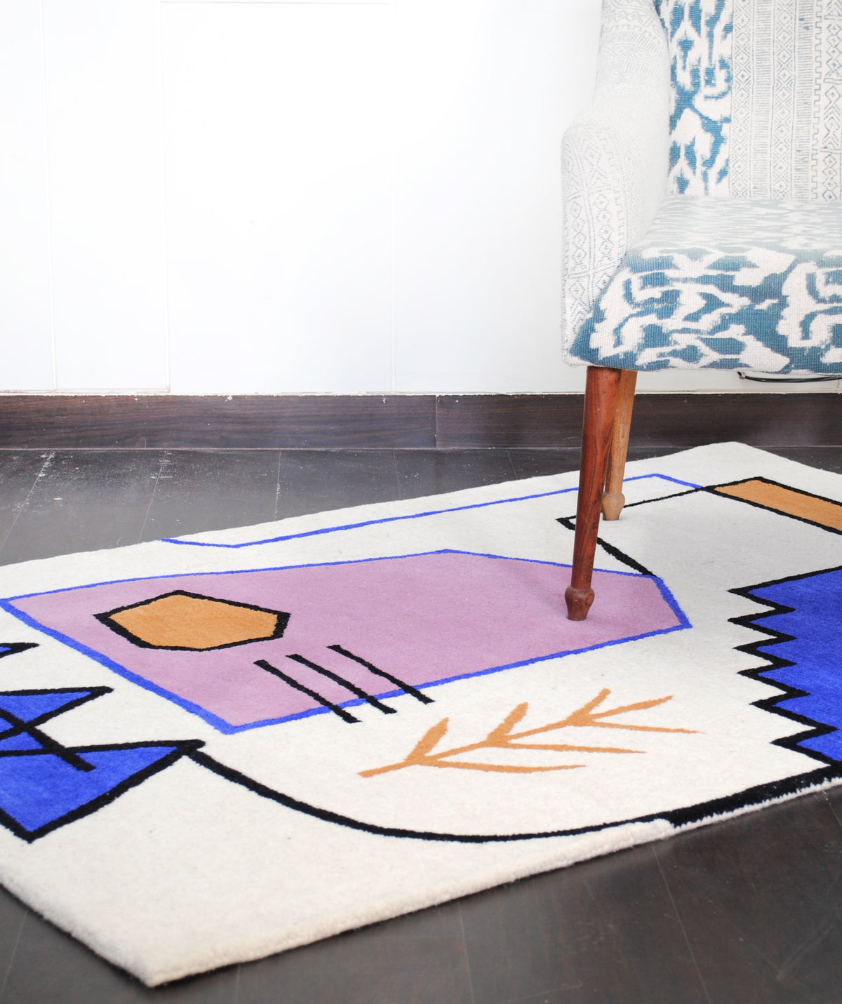 Hampi hand-tufted wool rug on a dark floor under an upholstered chair, the lilac gable, cobalt stepped form and ochre sprig on an ivory ground, Art Avenue