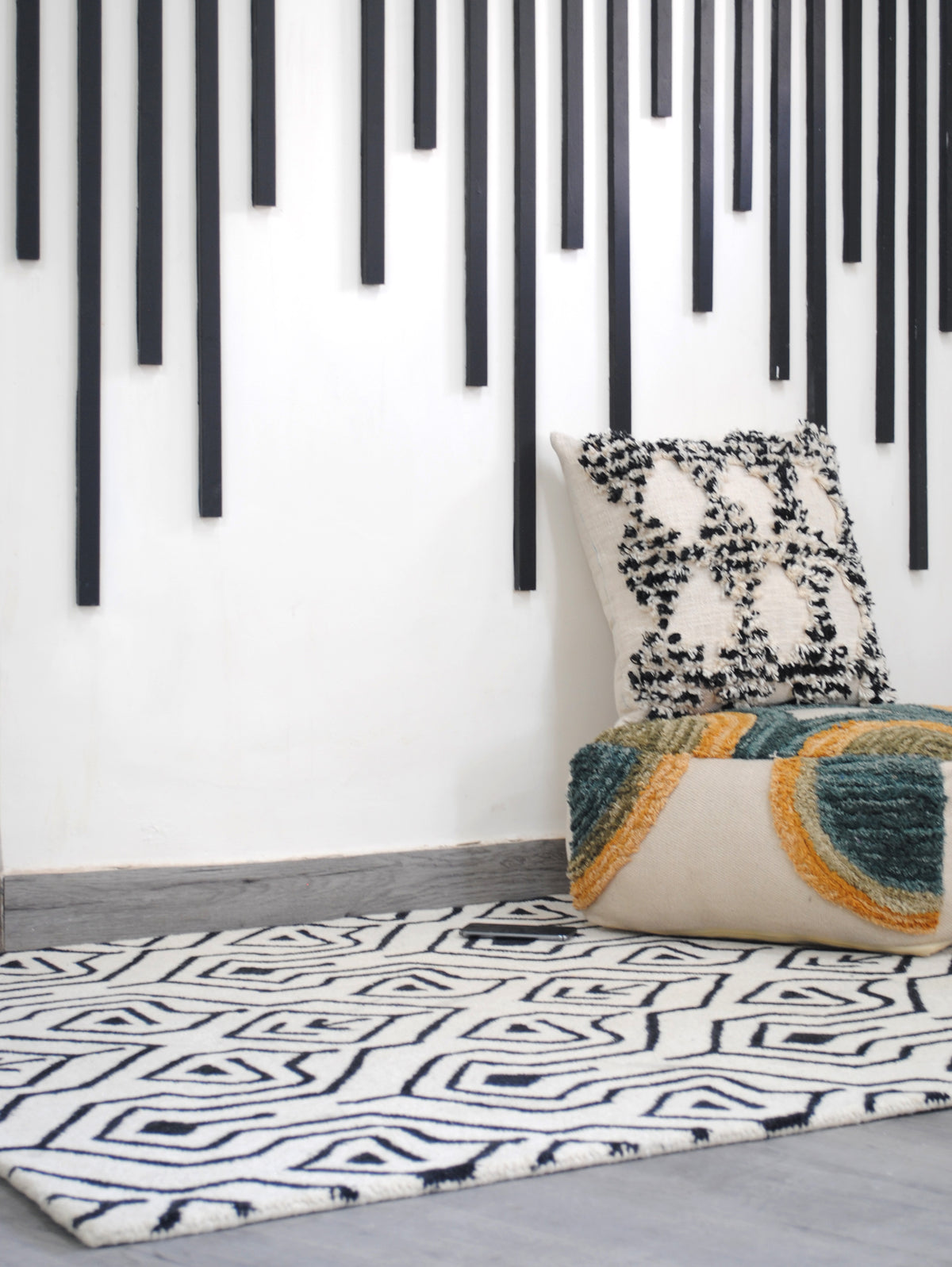 Chettinad hand-tufted rug in ivory and black styled in a white room beneath a black batten wall installation, with a tufted cushion and floor pouf, Art Avenue