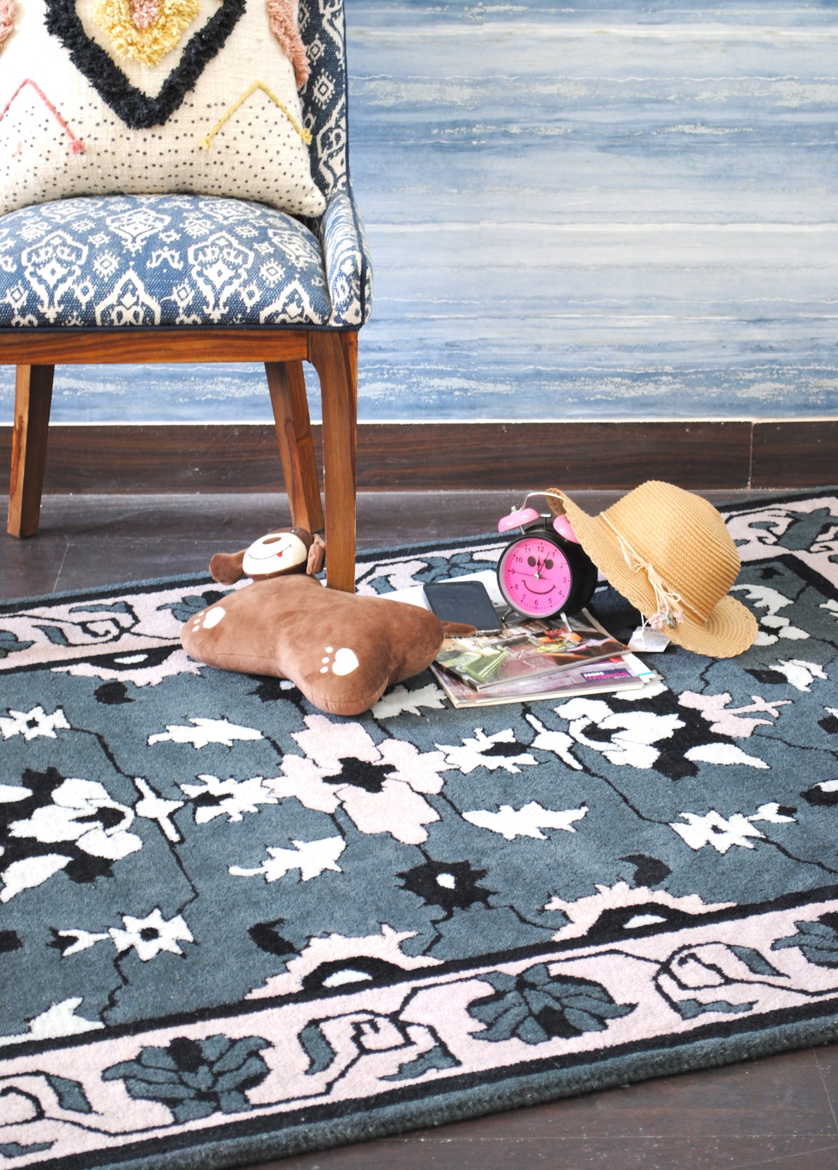 Shimla floral wool rug under an ikat upholstered chair on a dark wood floor, with a straw hat and pink clock