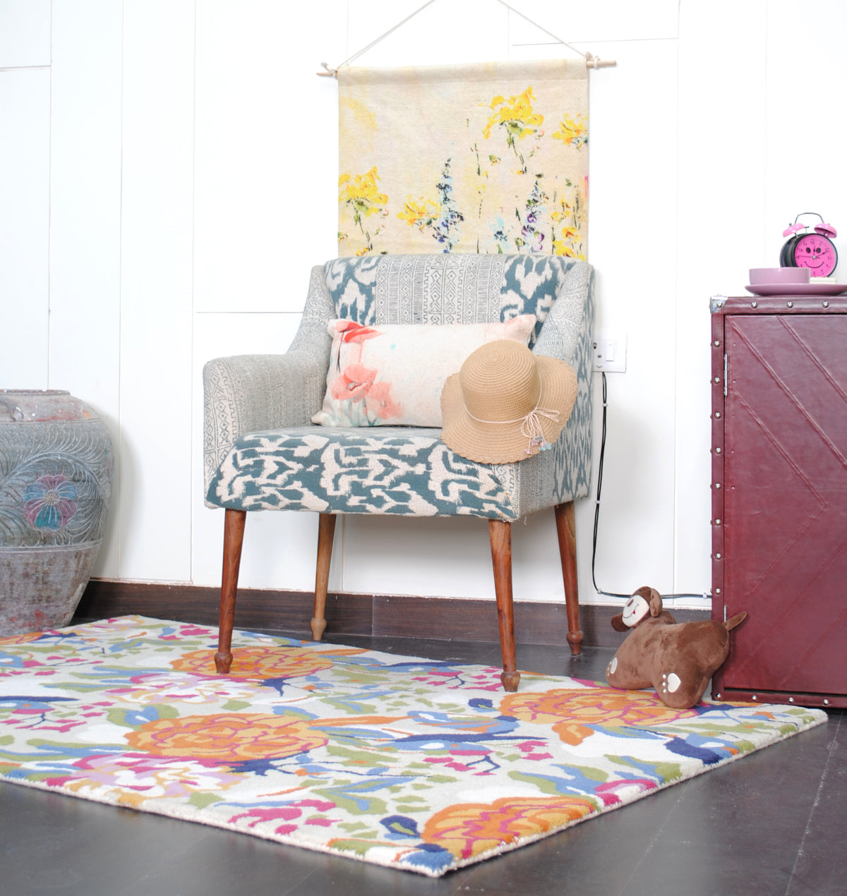 Ooty floral wool rug laid in front of an ikat armchair and a burgundy trunk, with a yellow botanical wall hanging behind