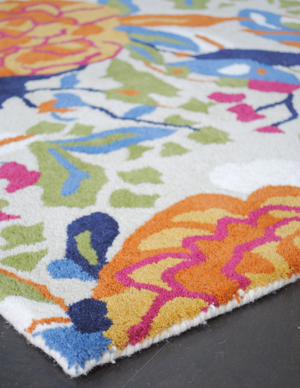 Corner of the Ooty hand-tufted rug: an orange and marigold peony traced in fuchsia beside navy and blue birds, finished with an ivory wool-bound edge