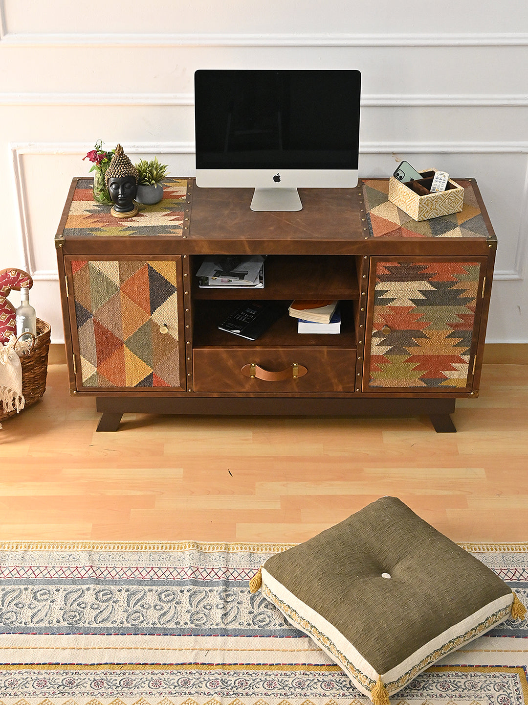 Mukundgarh kilim and leather TV unit at a three-quarter angle with a desktop screen, Buddha head and plants on top, beside a kilim pouf and cream armchair
