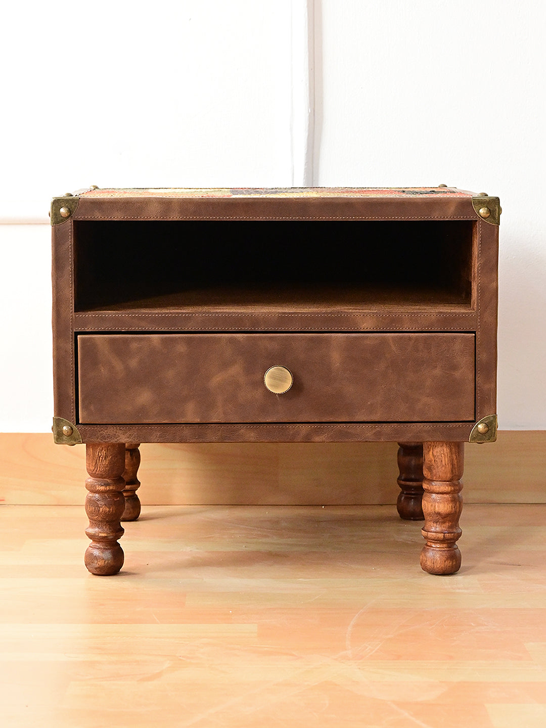 Front view of the Mahansar bedside table: leather front, open shelf over a drawer, brass corner caps