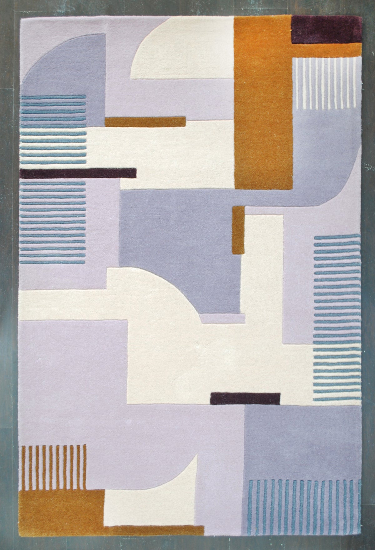 Mumbai hand-tufted wool rug laid flat on a dark wood floor, its lilac, ivory and ochre Art Deco composition seen from directly above, Art Avenue