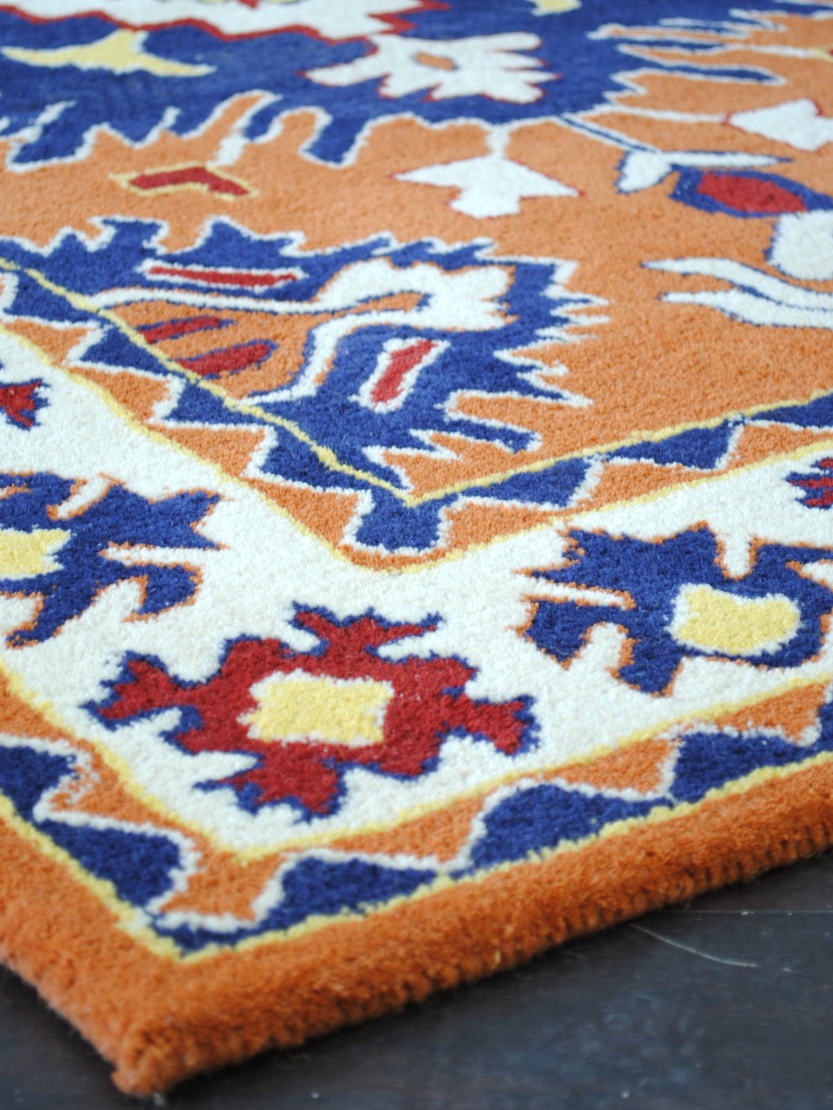 Corner of the Kutch hand-tufted wool rug, showing the ivory floral border, the saffron wool-bound edge and the dense even-cut pile, Art Avenue