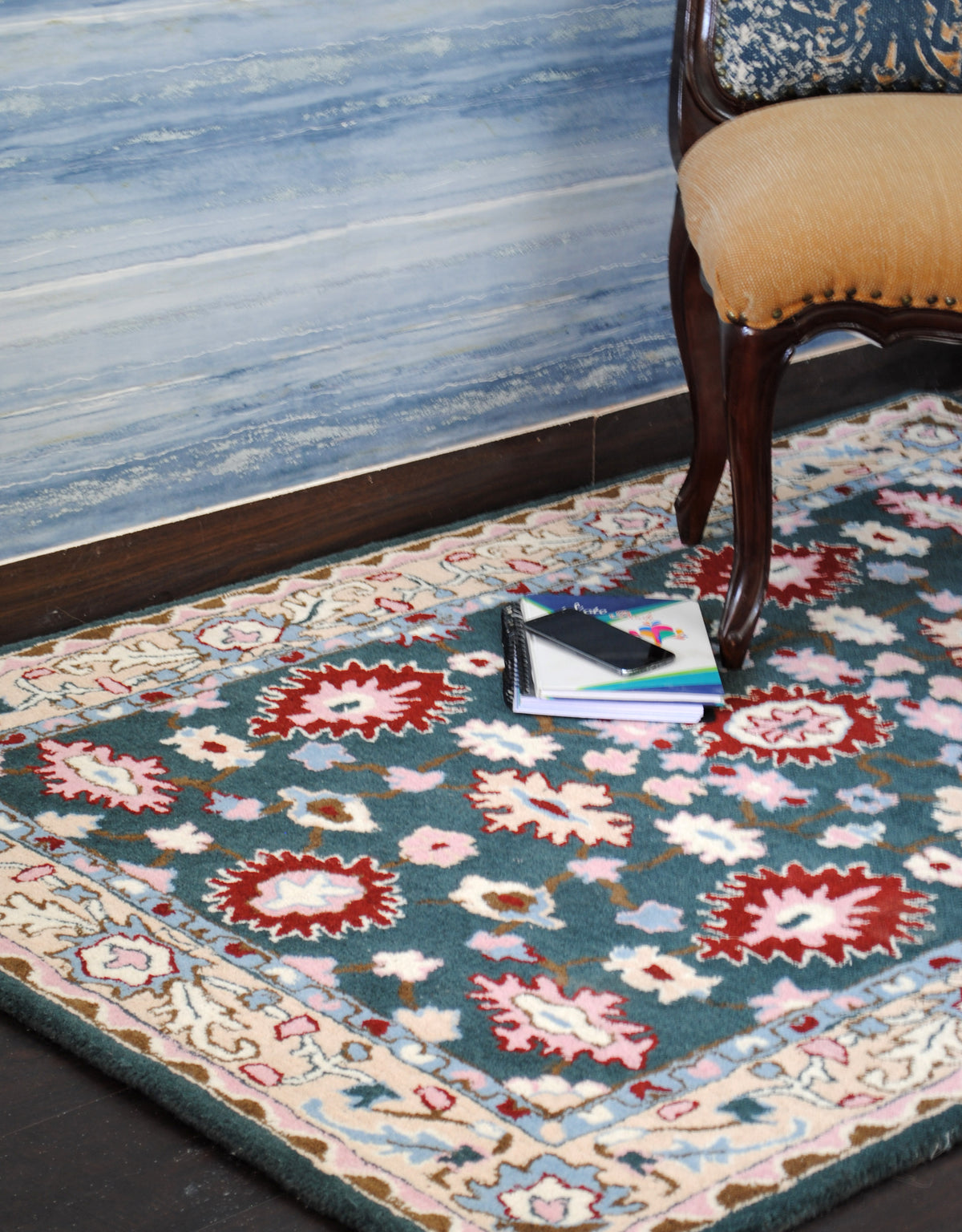 Srinagar teal floral wool rug in a room beside an upholstered wooden chair, cream border and deep teal field in view