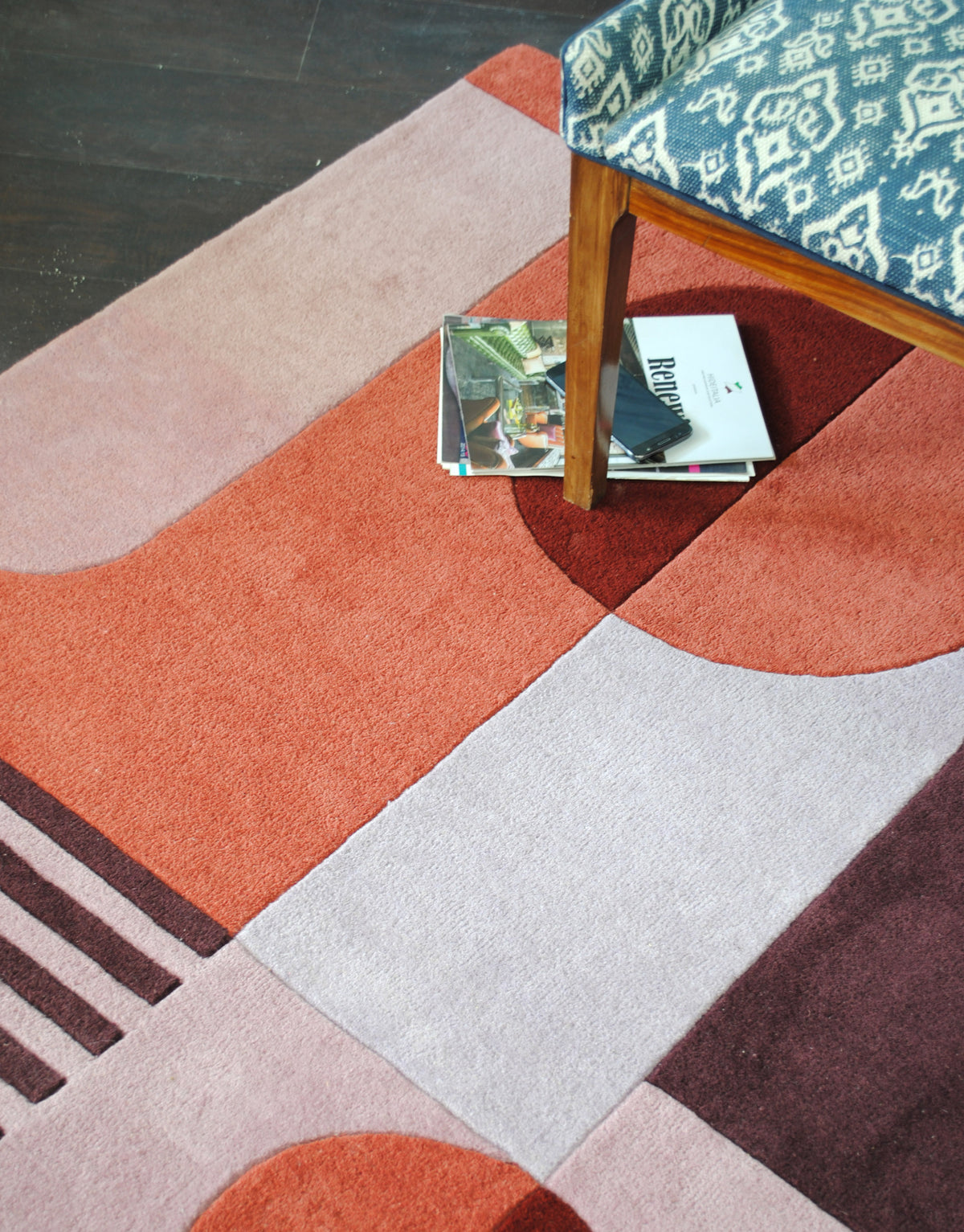 Angled view of the Konark rug under an upholstered chair, lilac grey and terracotta blocks with plum stripes