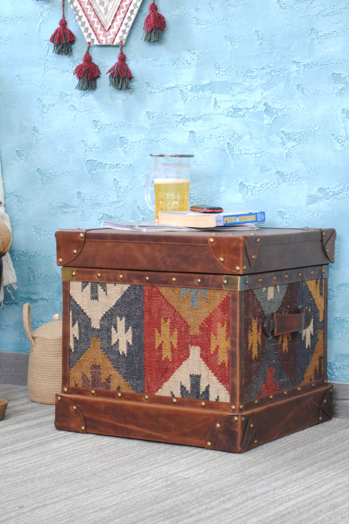 Madras leather-top kilim trunk styled as a side table against a blue wall, books and a glass on the leather lid