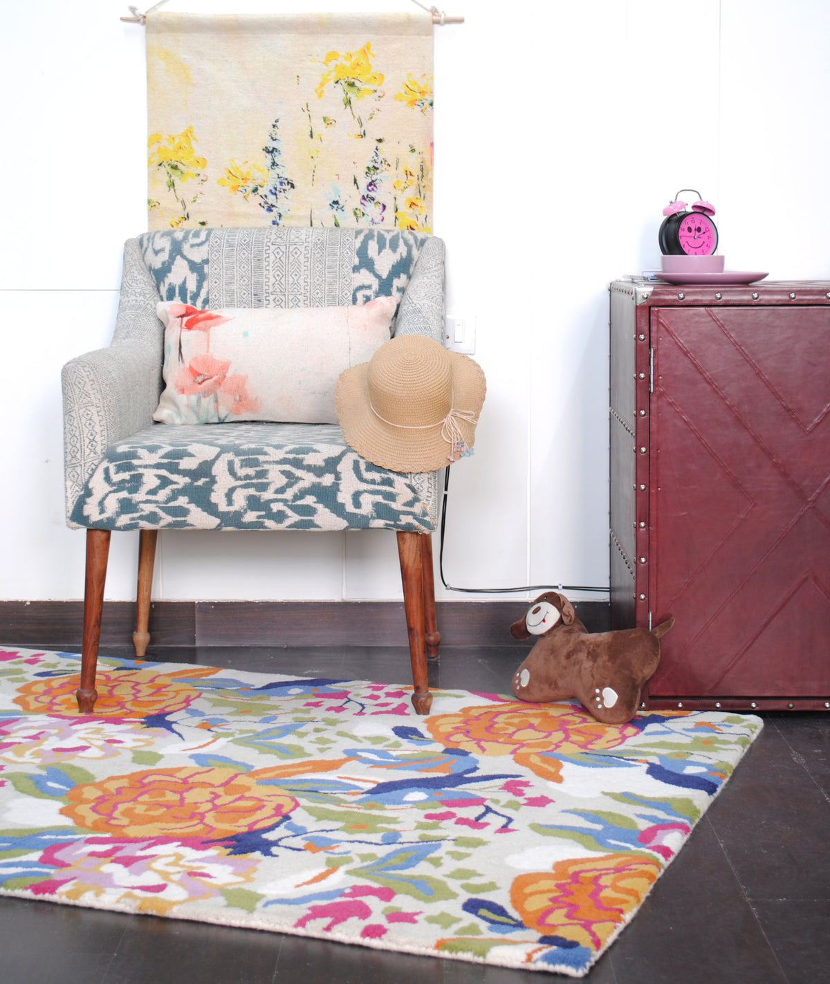 Ooty hand-tufted rug in a portrait room view: an ikat armchair, a botanical wall hanging and a burgundy trunk above the orange and blue floral rug