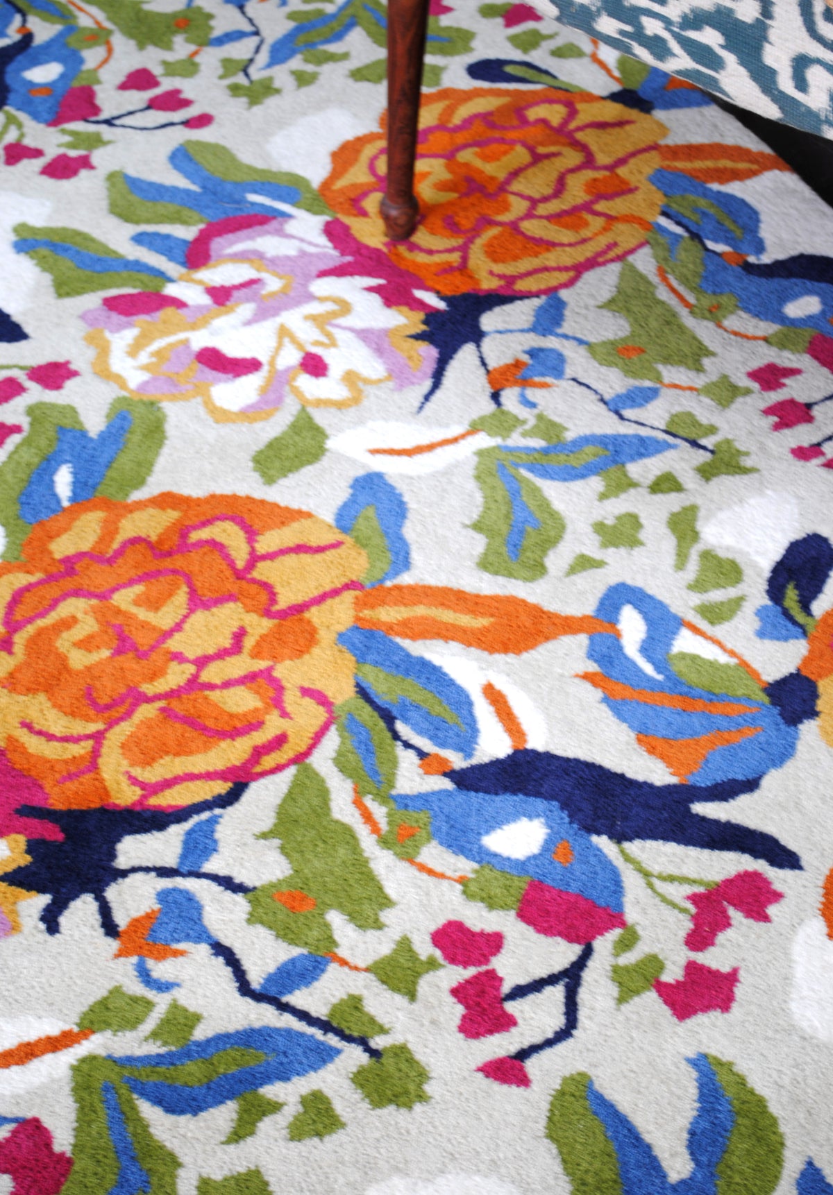 Top-down detail of the Ooty rug: an orange peony, a lilac and white bloom and navy birds among olive leaves, beside a turned wooden chair leg
