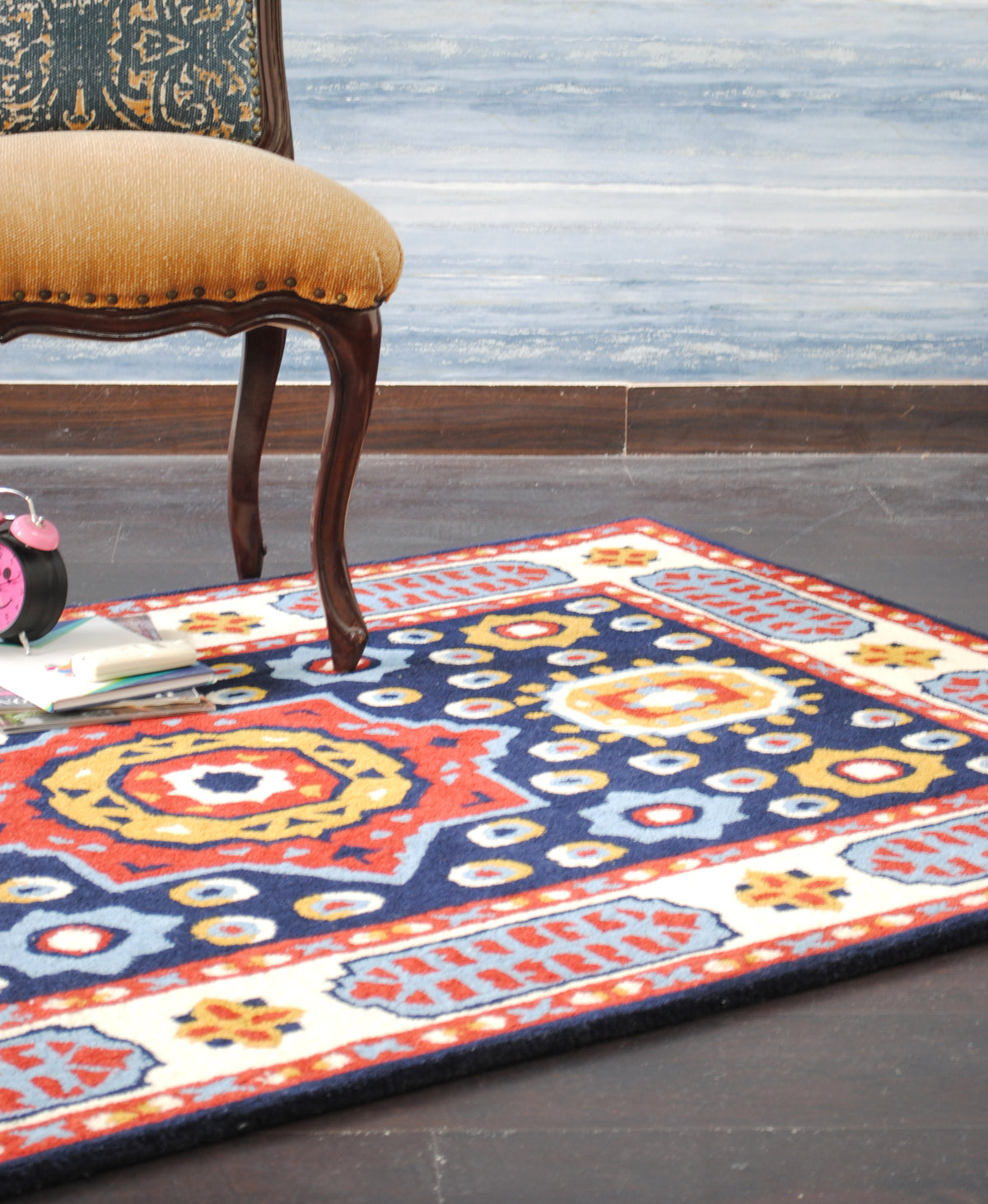 Orchha hand-tufted rug on a dark floor beside a carved chair, ivory border and red guard stripes in view