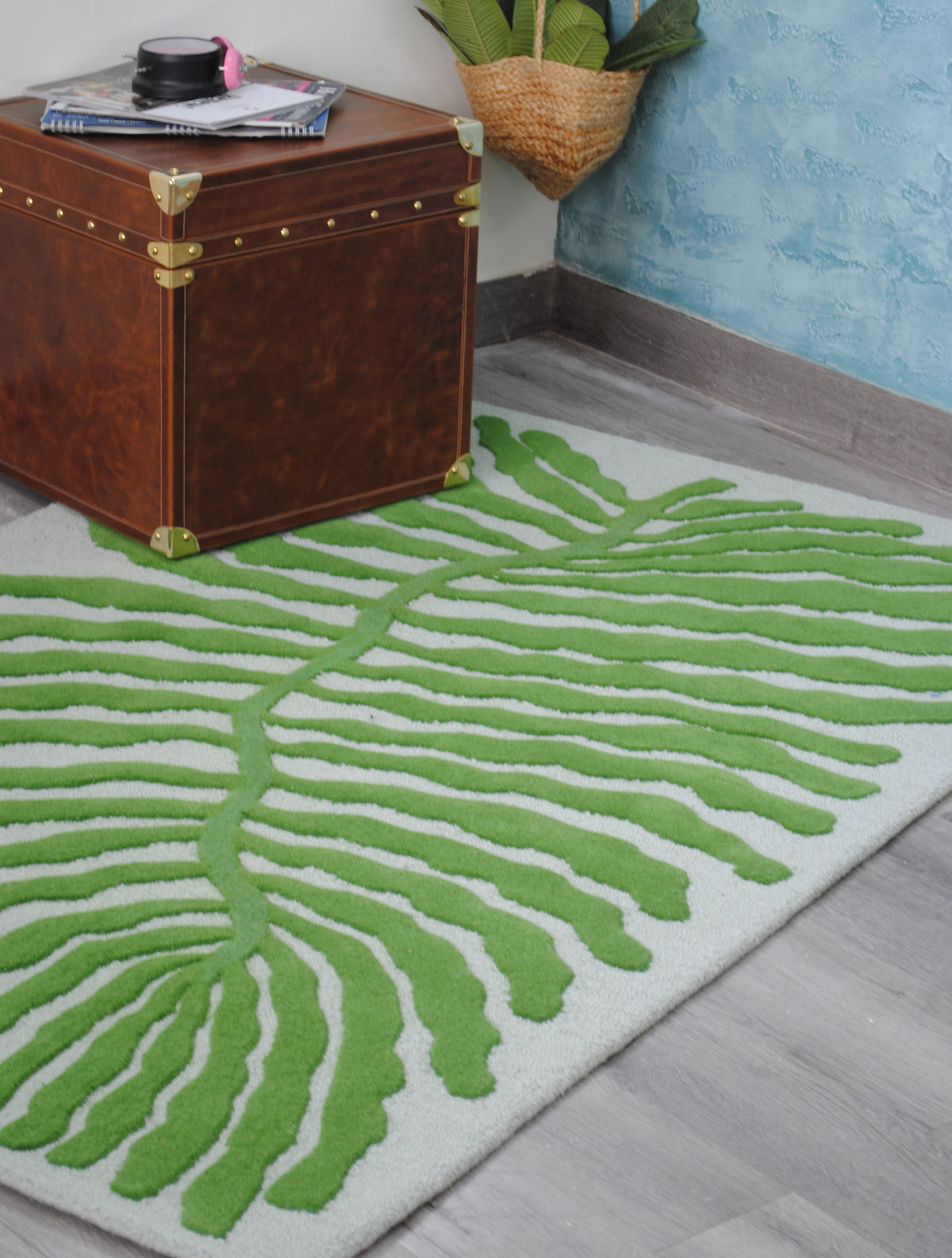 Wider view of the Munnar rug's winding green midrib and fronds beside a brown leather cube trunk