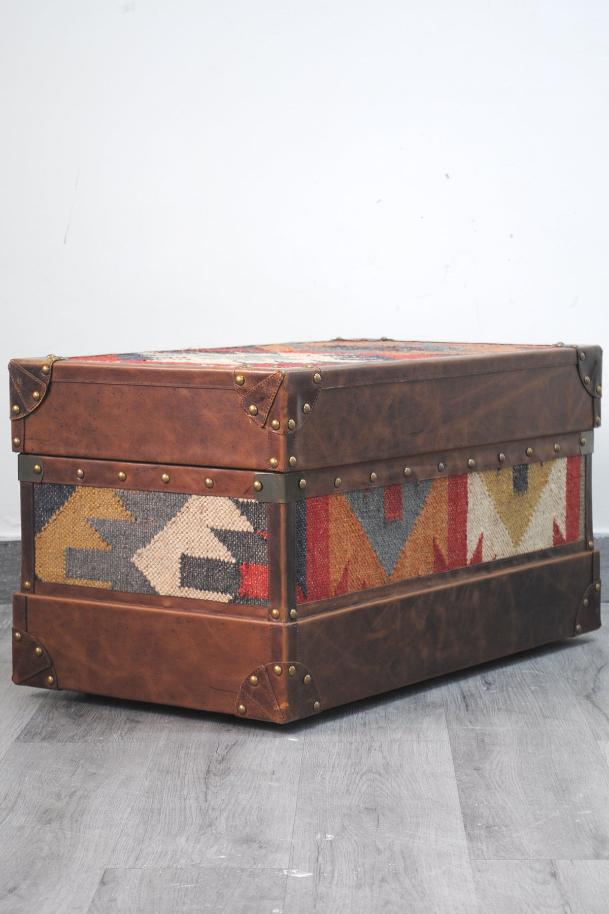 Angled view of the Bundi kilim steamer trunk showing the kilim lid, side panel and brass-studded leather bands