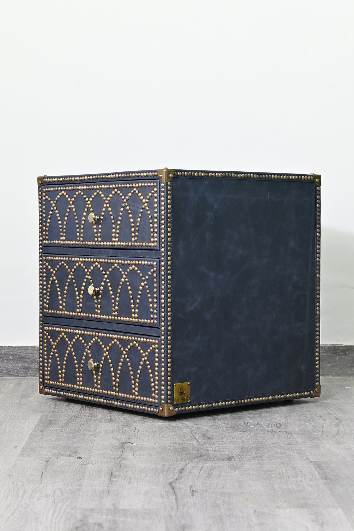 Samode studded leather bedside chest at a three-quarter angle, showing the brass nailhead drawer fronts and plain navy leather flank