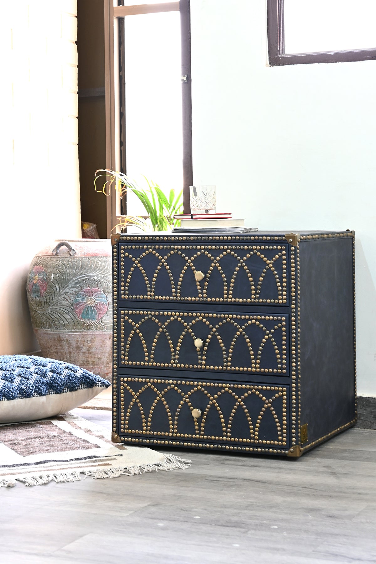 Samode studded leather bedside chest in a styled corner with a carved urn and woven rug