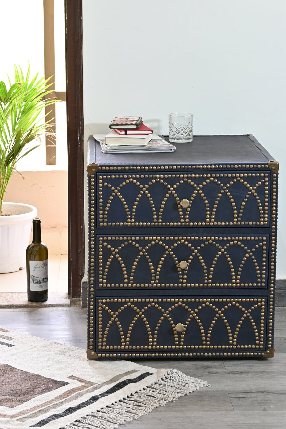 Samode studded leather bedside chest styled in a sunlit room with books and a wine bottle