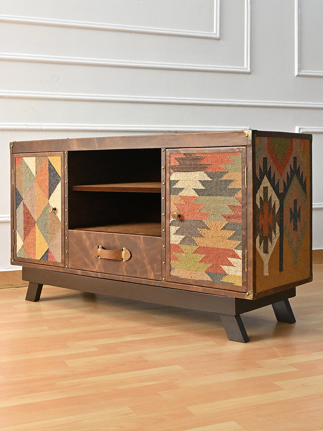 Mukundgarh TV unit at a three-quarter angle showing the full-height vintage kilim flank, studded leather binding and brass corner caps