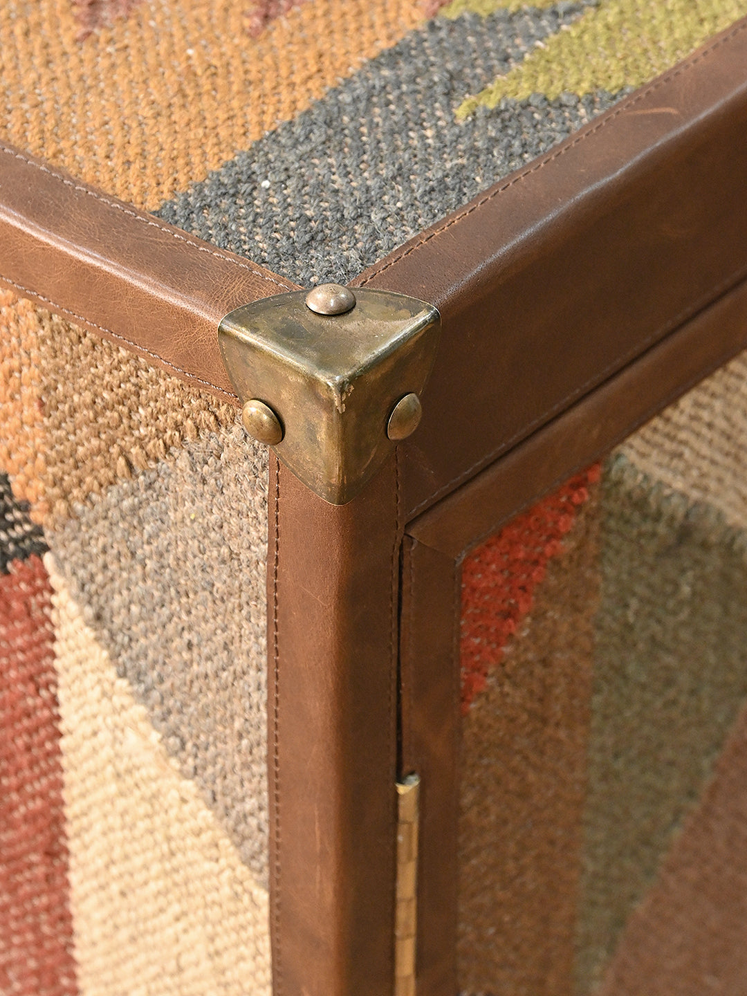 Close-up of a brass corner cap where the stitched leather binding meets the kilim top of the Mukundgarh TV unit
