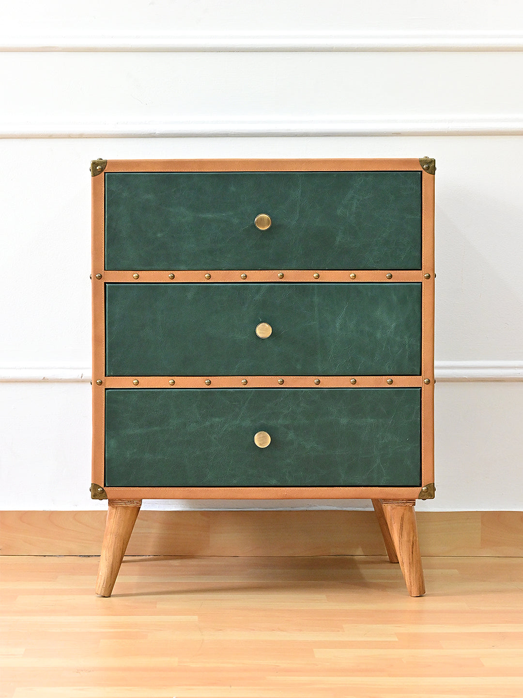 Sariska chest of drawers, front view — three bottle-green leather drawers with brass knobs, tan studded banding and tapered solid wood legs
