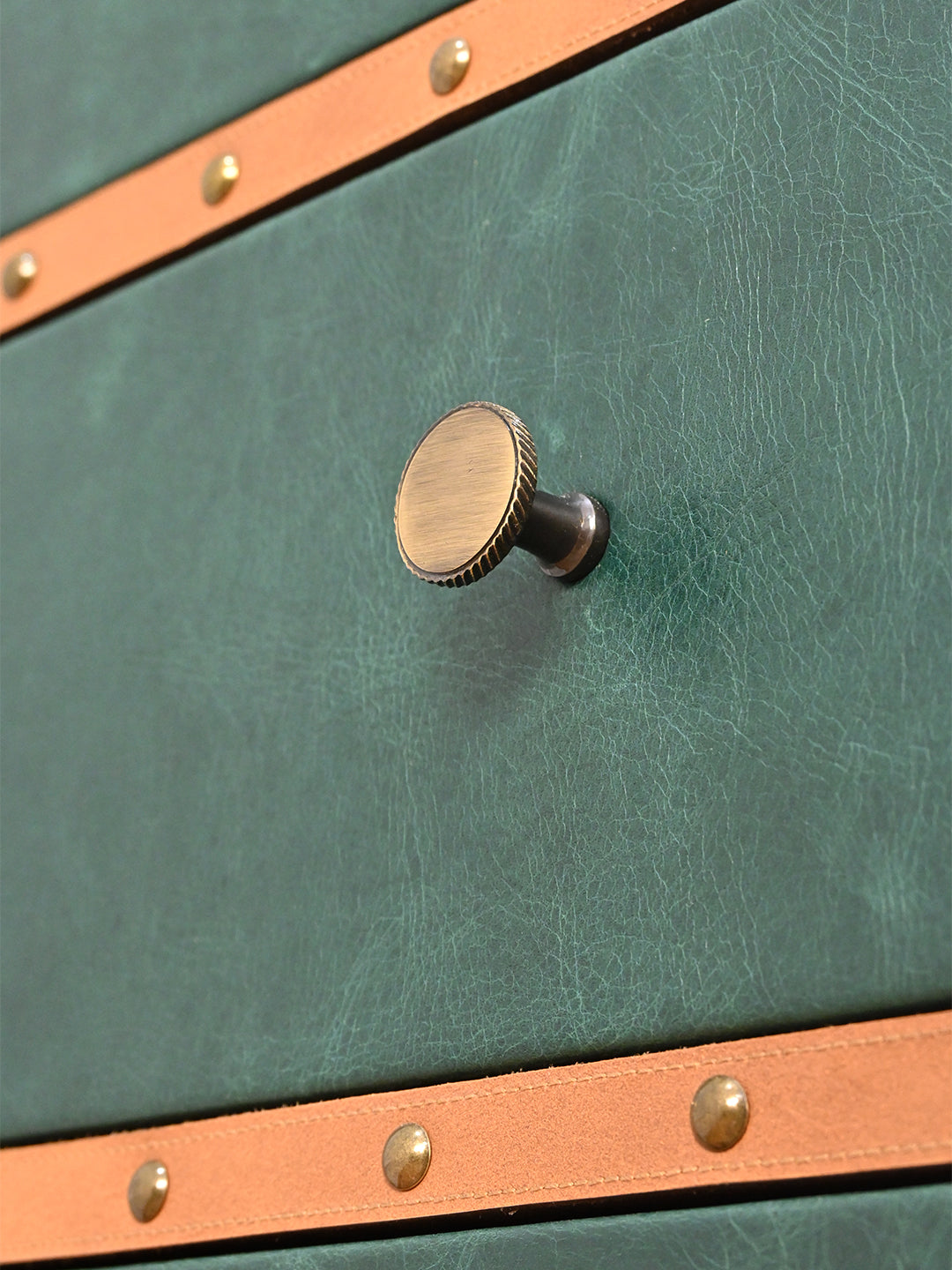 Knurled-edge antique brass knob on a green leather drawer front, between tan leather bands set with brass studs