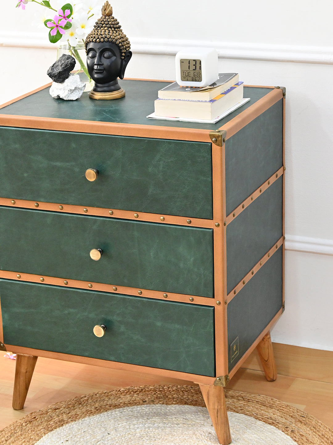 Sariska chest of drawers from above, a Buddha head, orchids, books and a clock on the green leather top