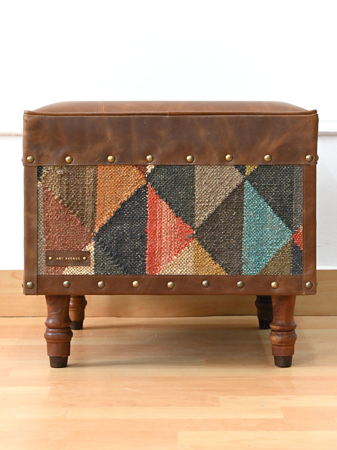 Front view of the Dundlod ottoman by Art Avenue, geometric kilim panel framed in studded leather on turned wooden legs