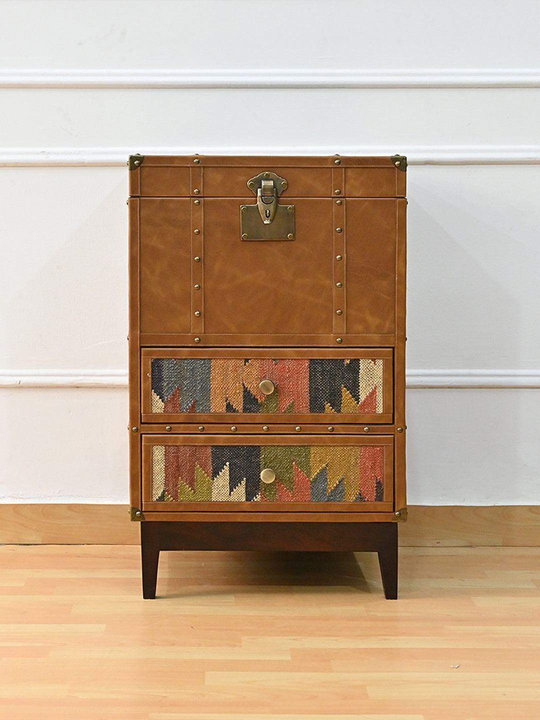 Osian drawer trunk seen straight on — tan leather lid and front with a brass lock plate above two vintage kilim drawer fronts, on dark tapered legs. Art Avenue, made to order.