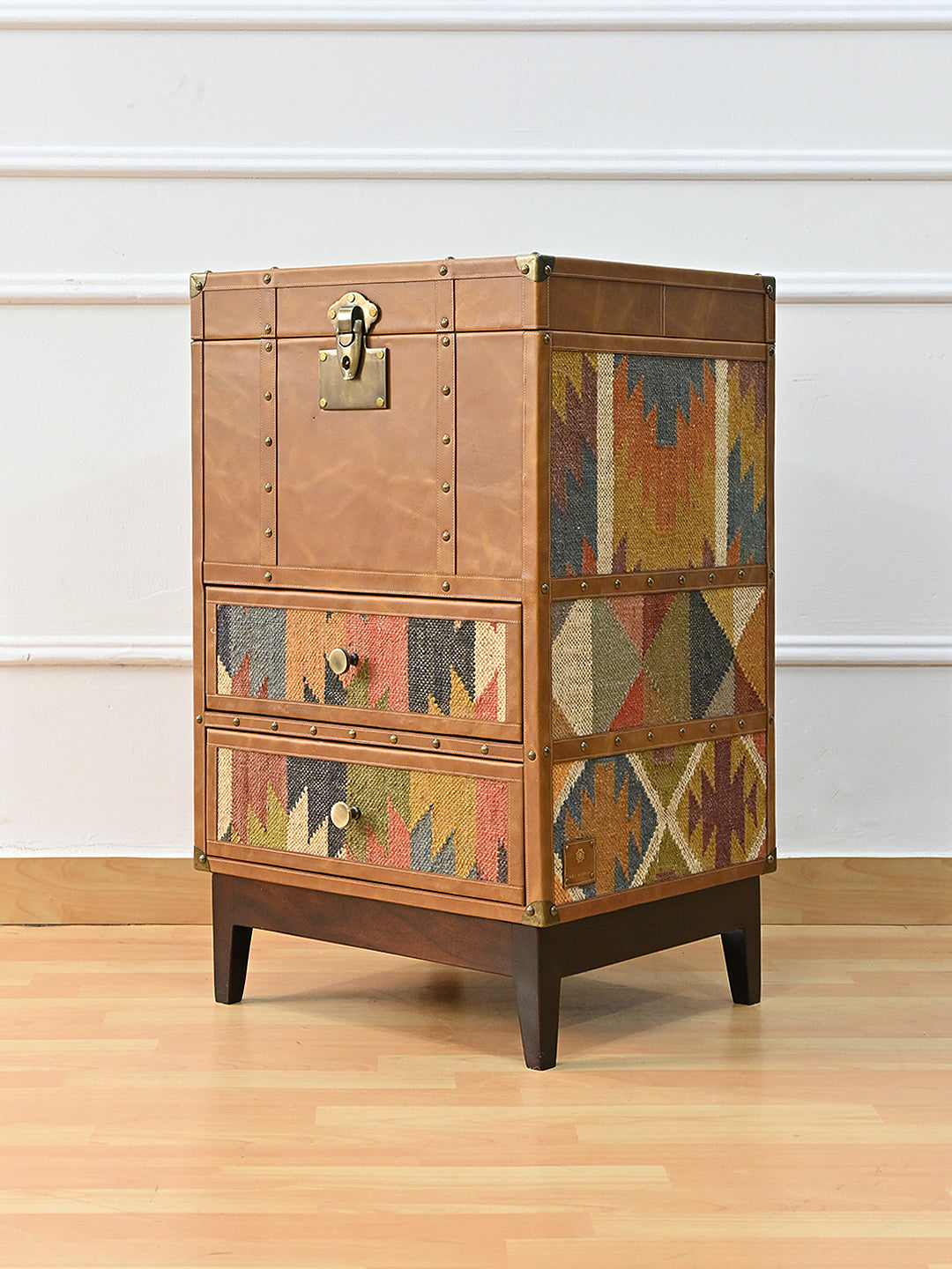 Three-quarter view of the Osian trunk showing the studded tan leather front beside a full-height vintage kilim flank, with brass corner caps. Art Avenue, made to order.